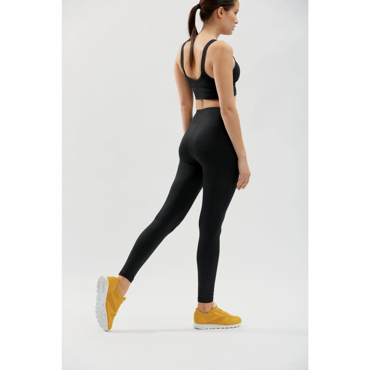 Black active leggings worn by a model, paired with a black sports top and yellow trainers, featuring a logo.