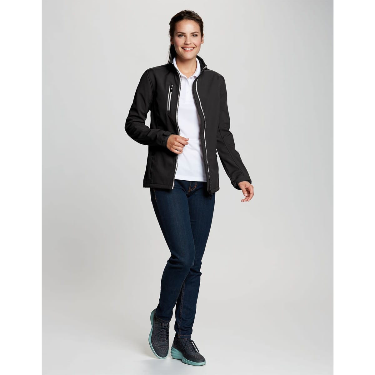 A woman wearing a black softshell jacket over a white top and jeans, featuring a logo on the jacket.