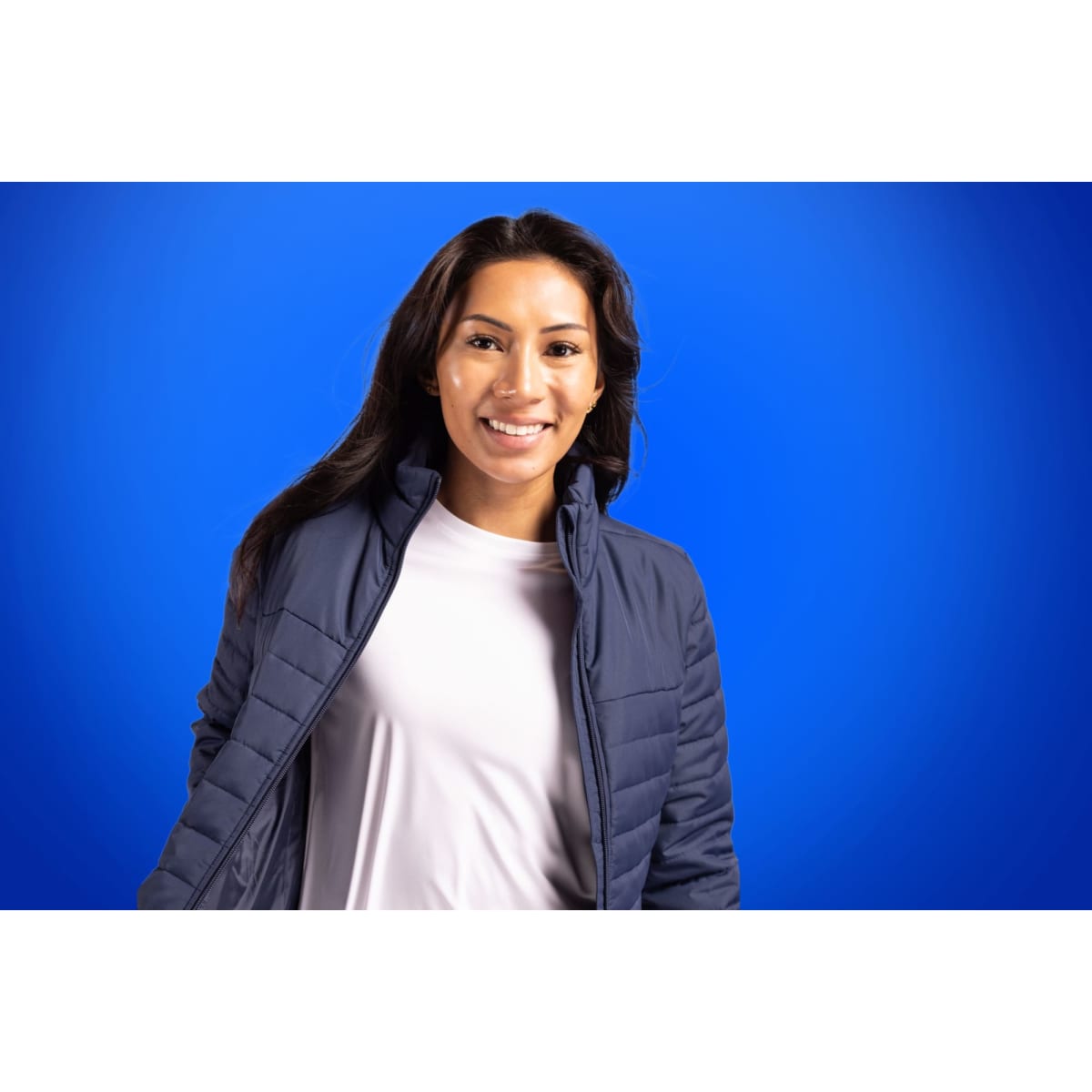 A woman wearing a navy puffer jacket over a light shirt, set against a blue background. The jacket has a logo.