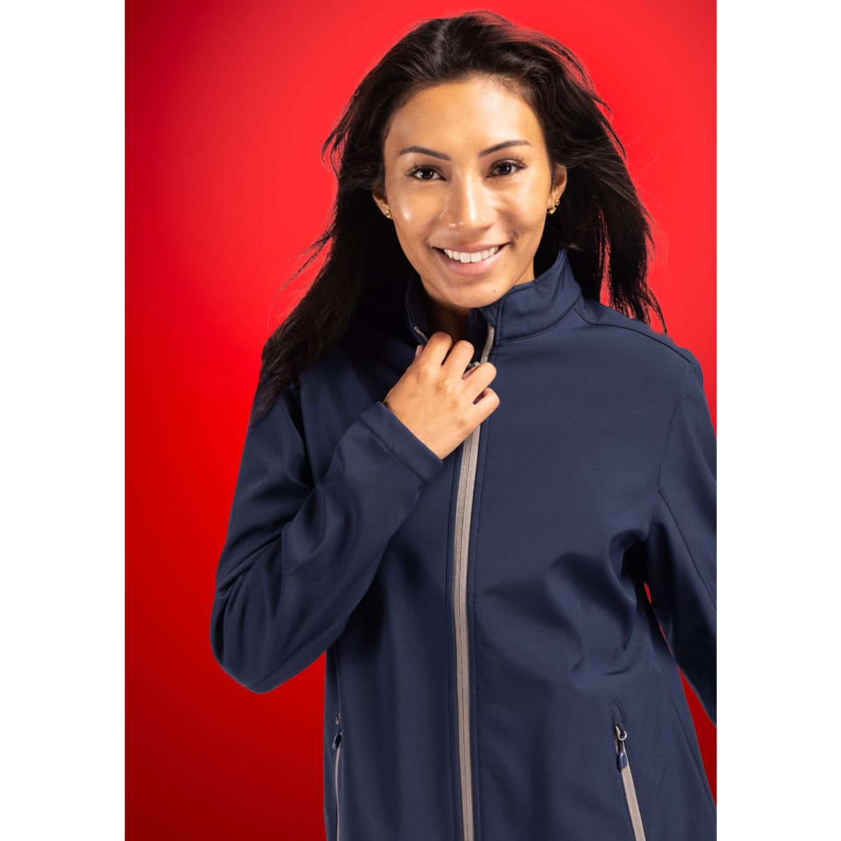 A woman smiles while wearing a navy soft shell jacket with a high collar and zip closure. The background is bright red.