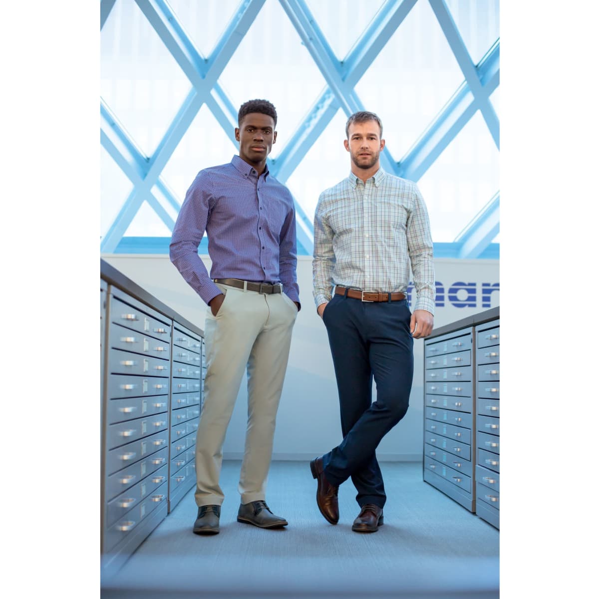 Two men standing in a bright space, wearing a patterned purple shirt and a plaid shirt with beige and navy pants.