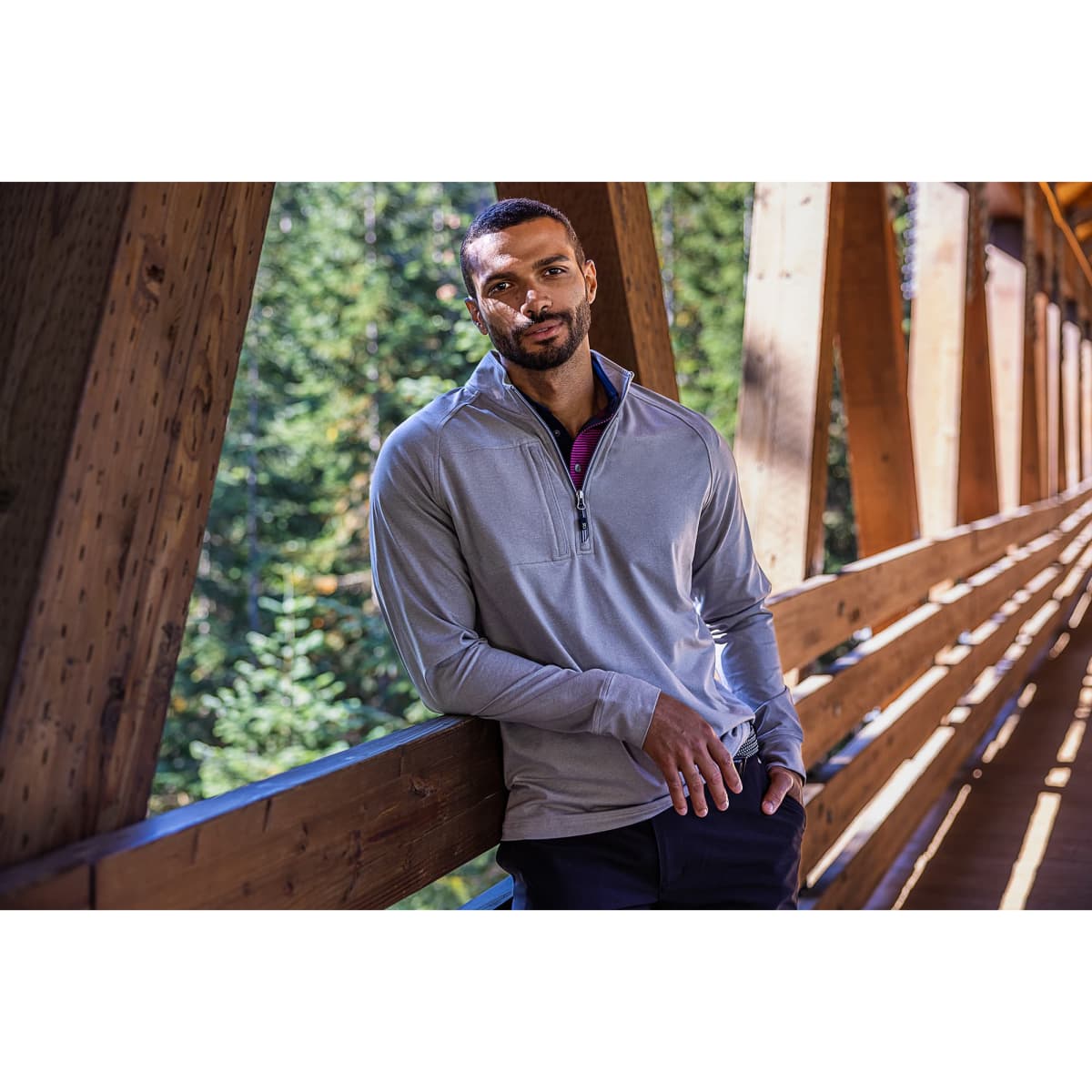 A grey half-zip jumper on a man leaning against a wooden railing, surrounded by trees. It features a logo.