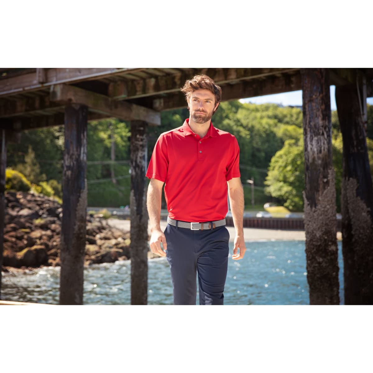 A man wearing a red polo shirt stands by a pier, surrounded by greenery and water. The shirt features a logo.