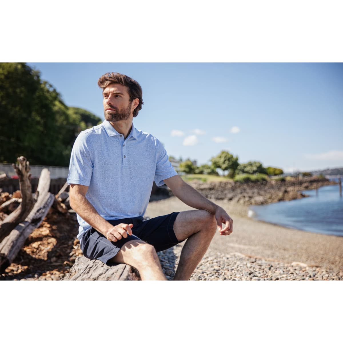 A man wearing a light blue botanical print polo shirt sits on a log by the water, set against a scenic background.