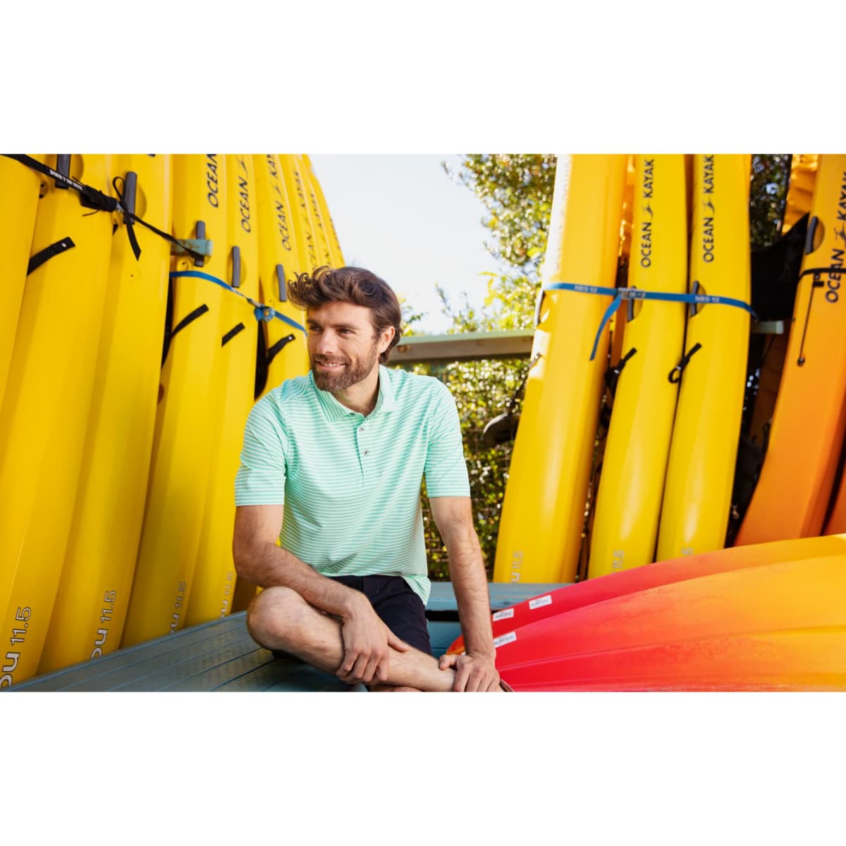 A man wearing a green and white striped polo shirt sits among bright yellow kayaks and colourful canoes.