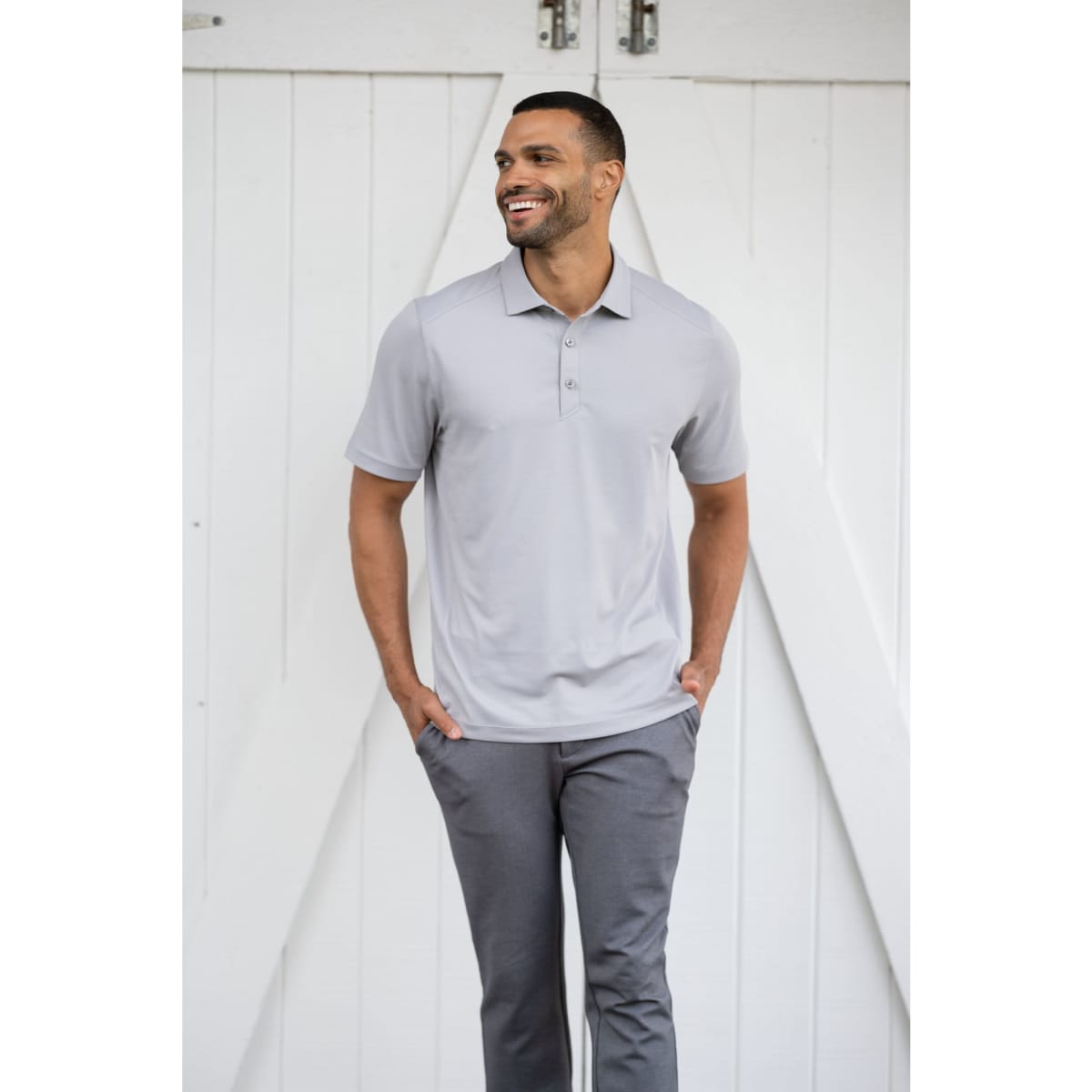 A man wearing a light grey polo shirt with a collar and buttons, paired with grey trousers, standing against a white backdrop.