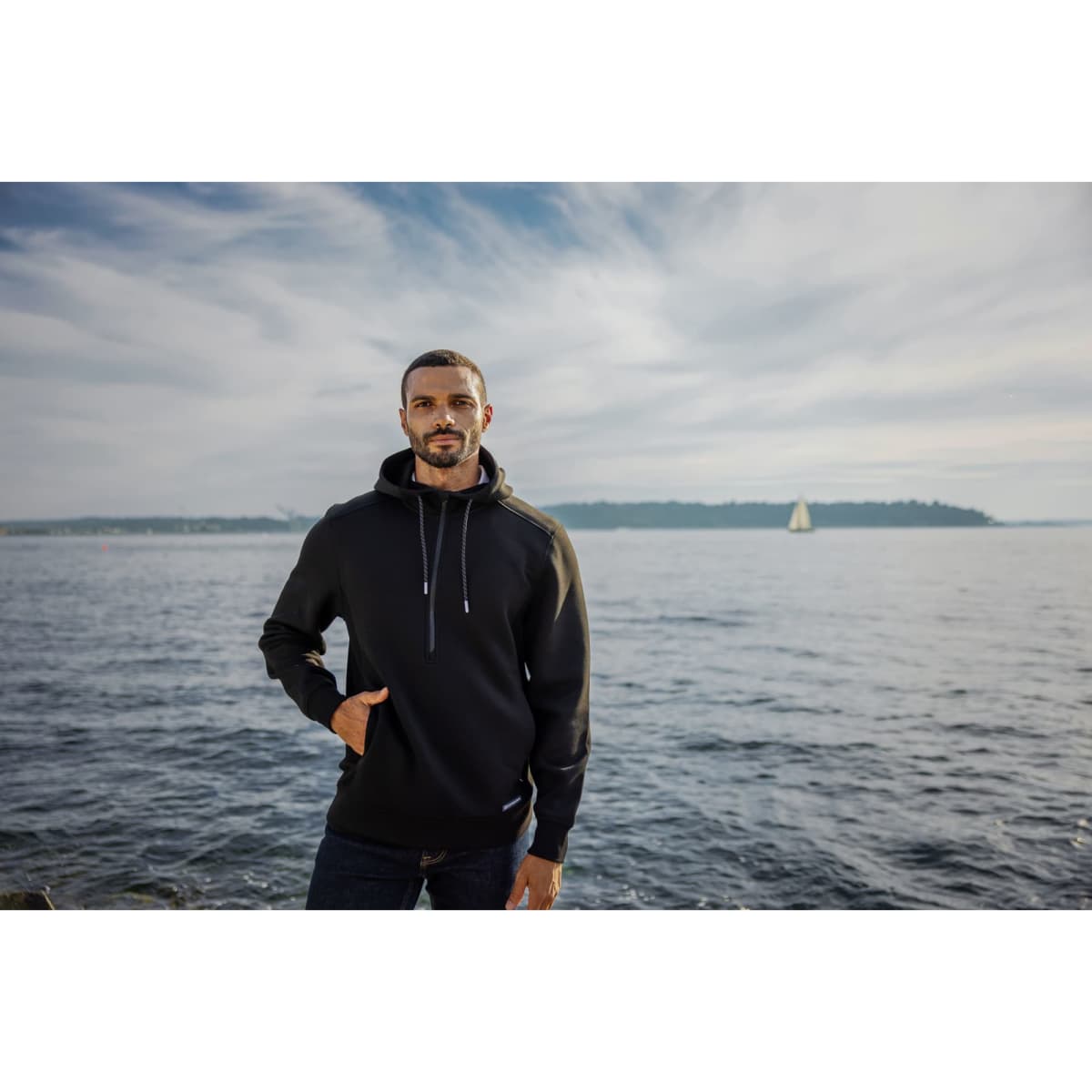 A man stands by the water wearing a black half zip hoodie, featuring a logo and a relaxed fit.