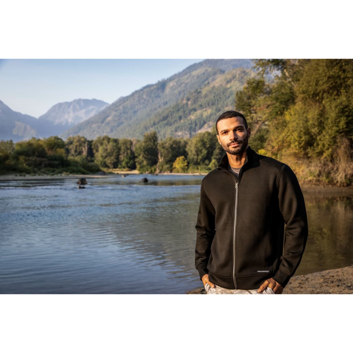 A man stands by a river wearing a black full zip jacket against a scenic mountain backdrop. The jacket features a logo.
