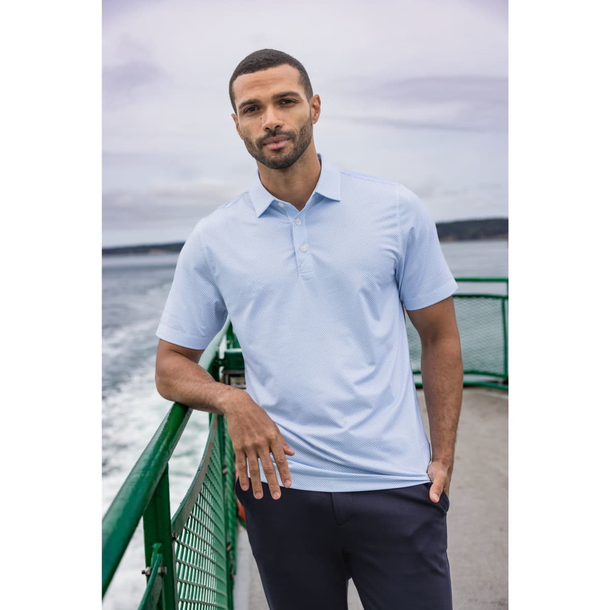 A light blue men's polo shirt features a subtle print and a logo, worn by a man on a ferry.