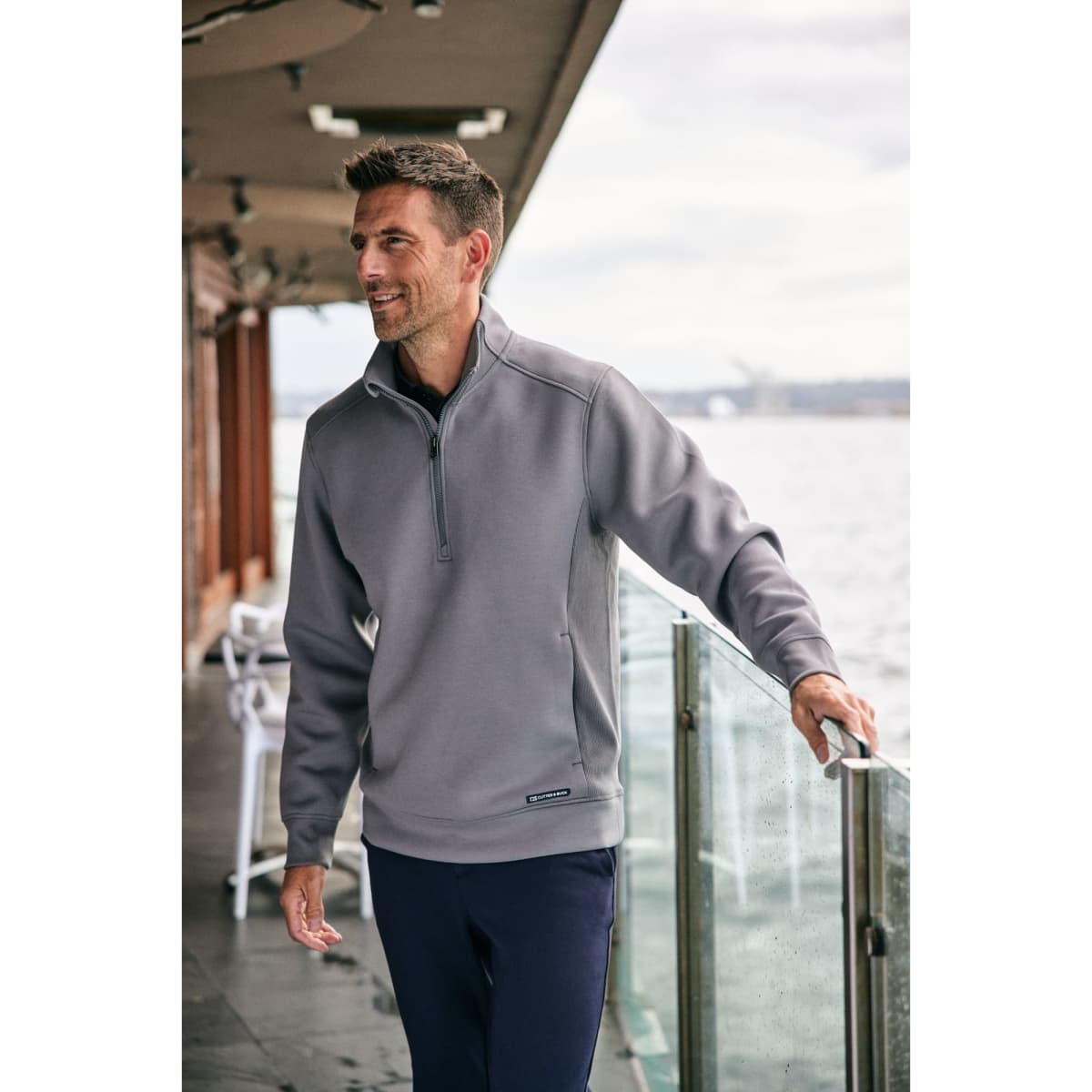A grey half zip jacket worn by a man standing near a railing by the water, featuring a logo.
