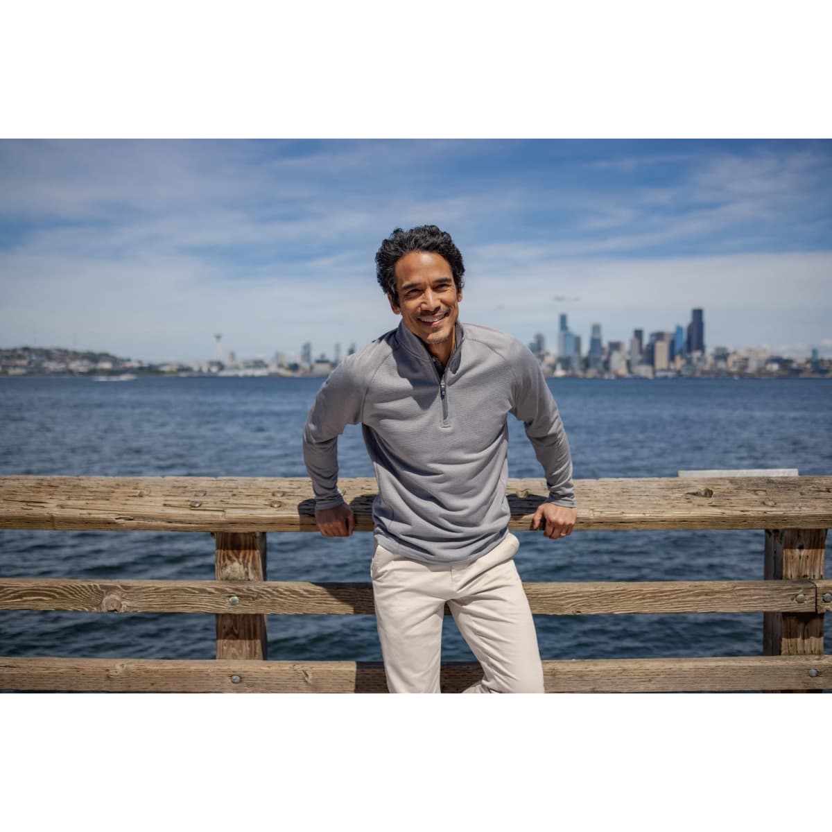 A man wears a light grey half-zip fleece jacket, standing by a wooden railing with a city skyline in the background.