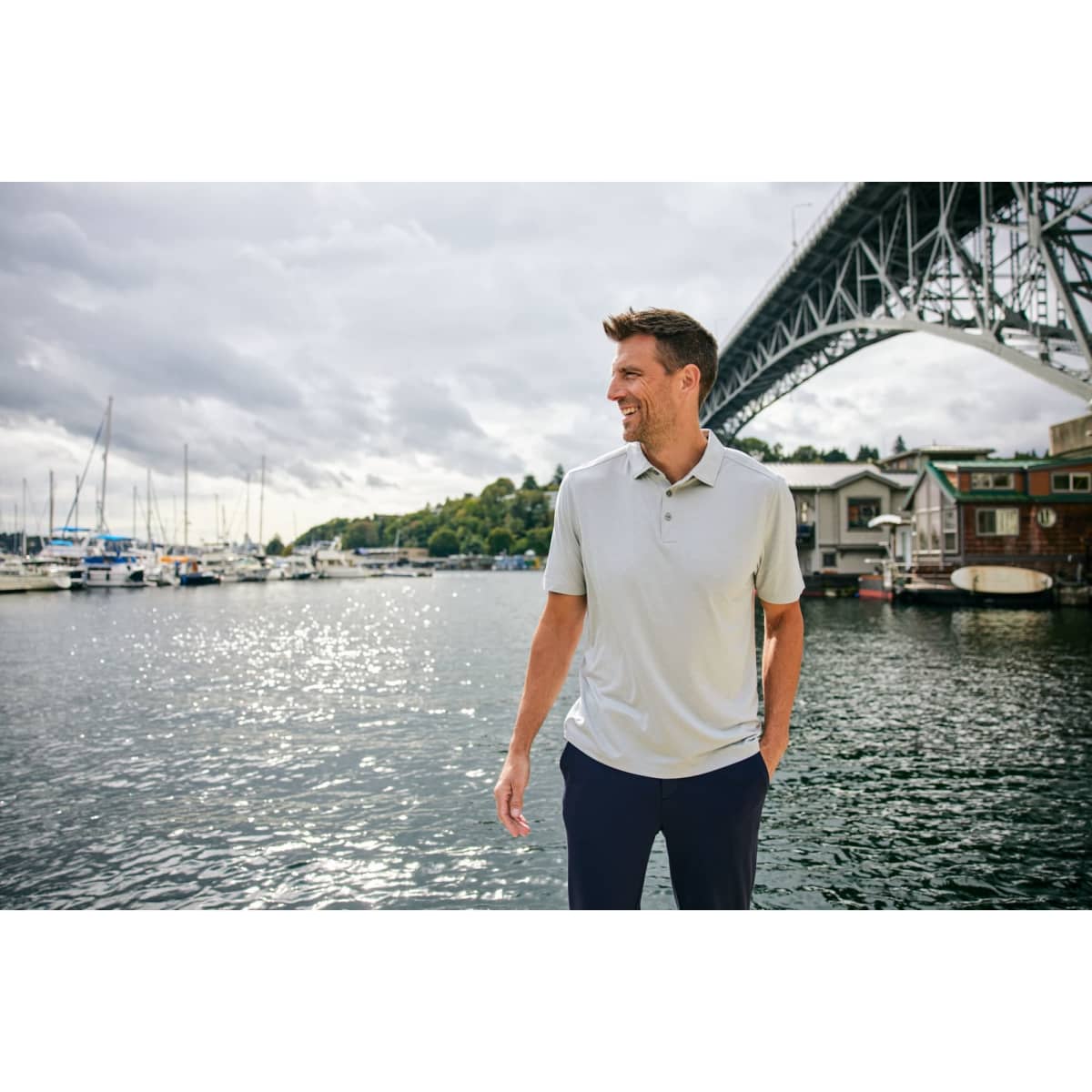 A man in a light grey polo shirt stands by the water, with boats and a bridge in the background.