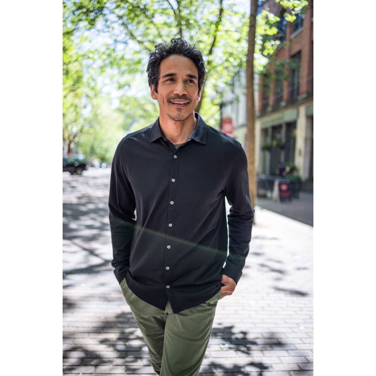 A long-sleeved, button-up shirt in black, modelled by a man outdoors in a tree-lined street.