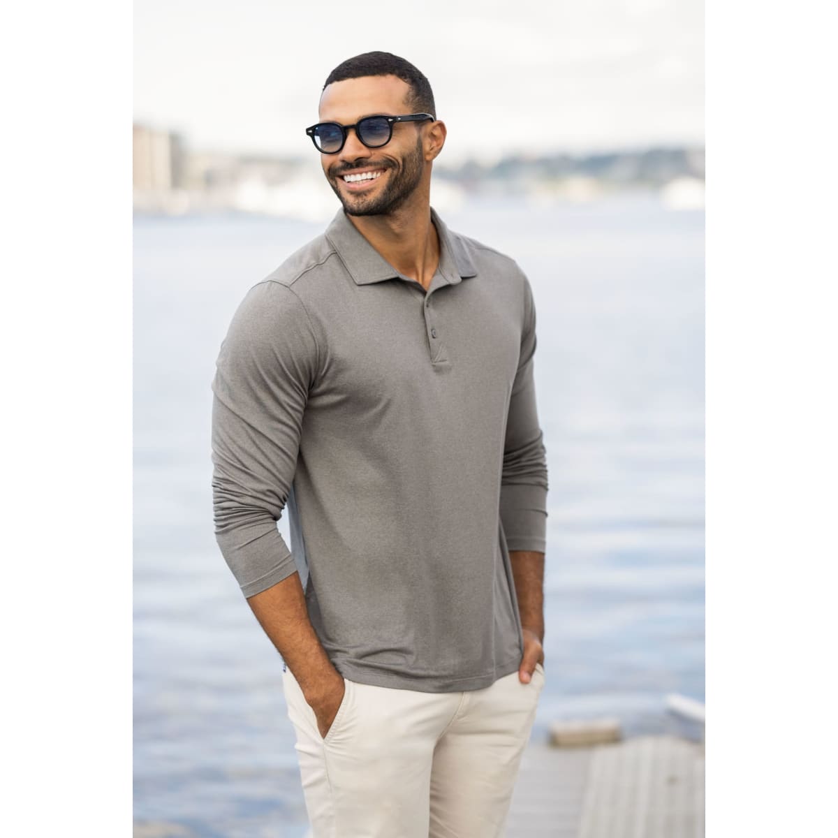 A man wearing a grey long sleeve polo shirt stands by the water, smiling, with sunglasses and light-coloured pants.
