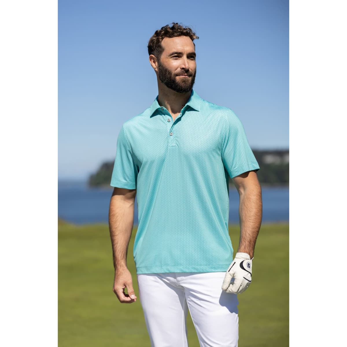 A man wearing a turquoise polo shirt stands outdoors, with a golf glove on his left hand and a scenic background.