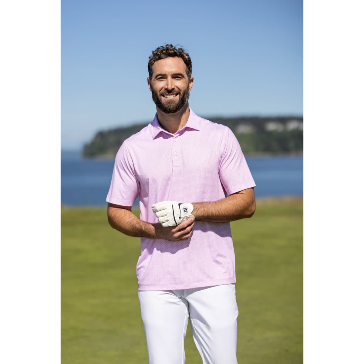 A man wearing a light pink polo shirt stands outdoors, with a golf glove on while posing against a scenic backdrop.