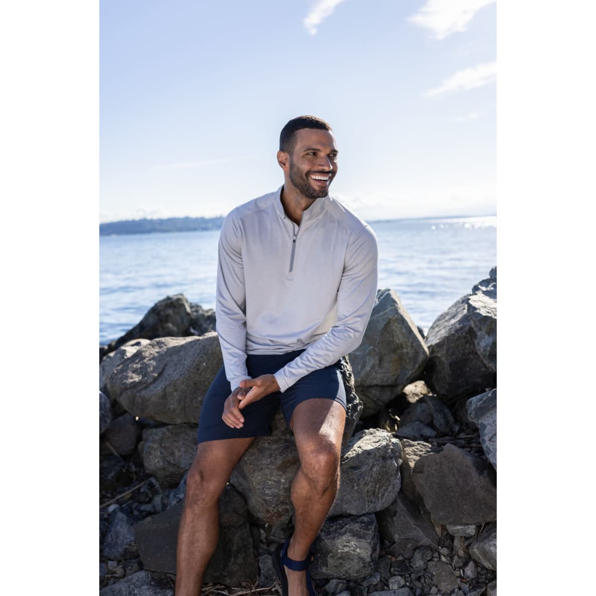 A man wearing a light grey quarter zip top sits on rocks by the water, smiling under a clear blue sky.