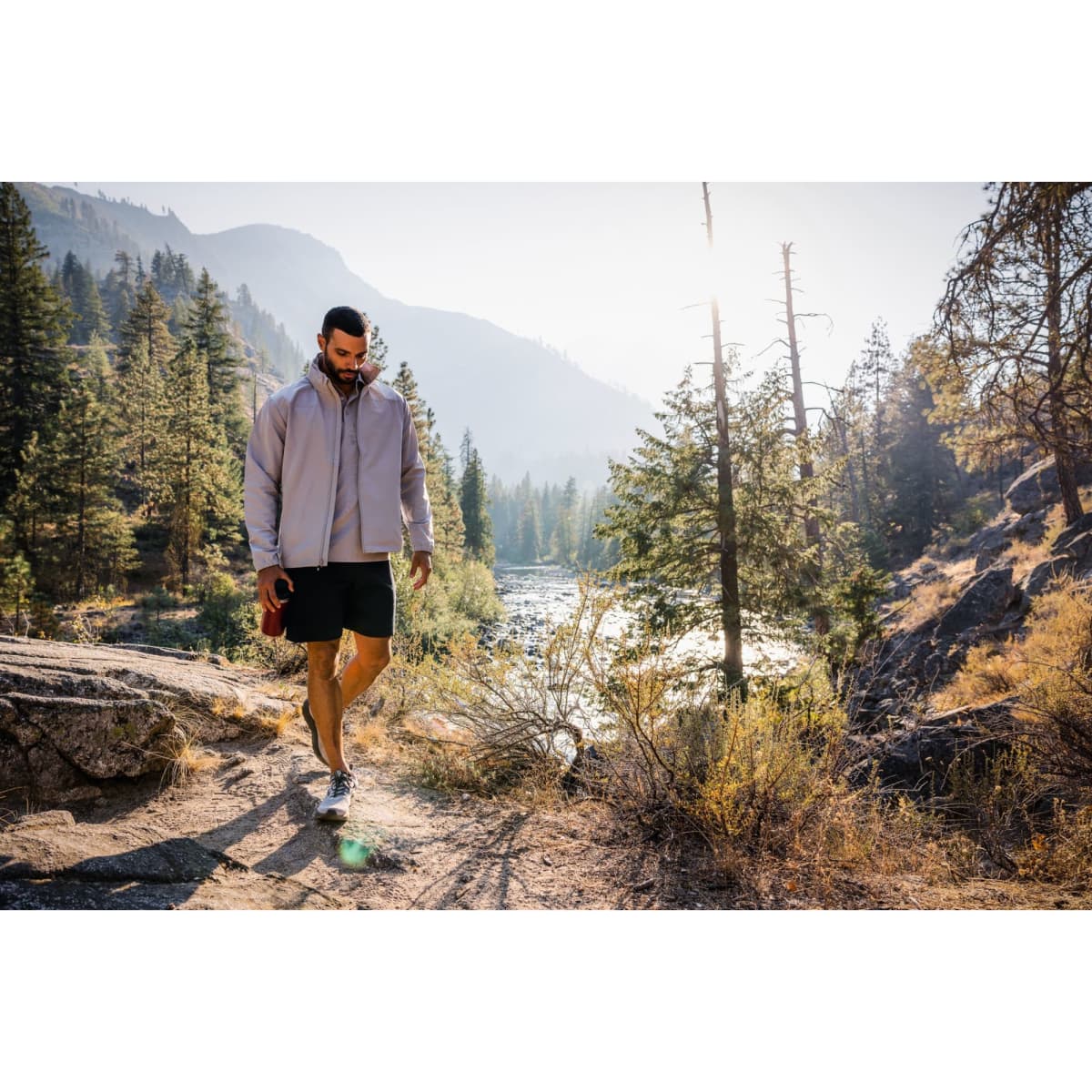 A man wearing a light grey full zip jacket and shorts walks along a rocky path near a river, surrounded by trees.