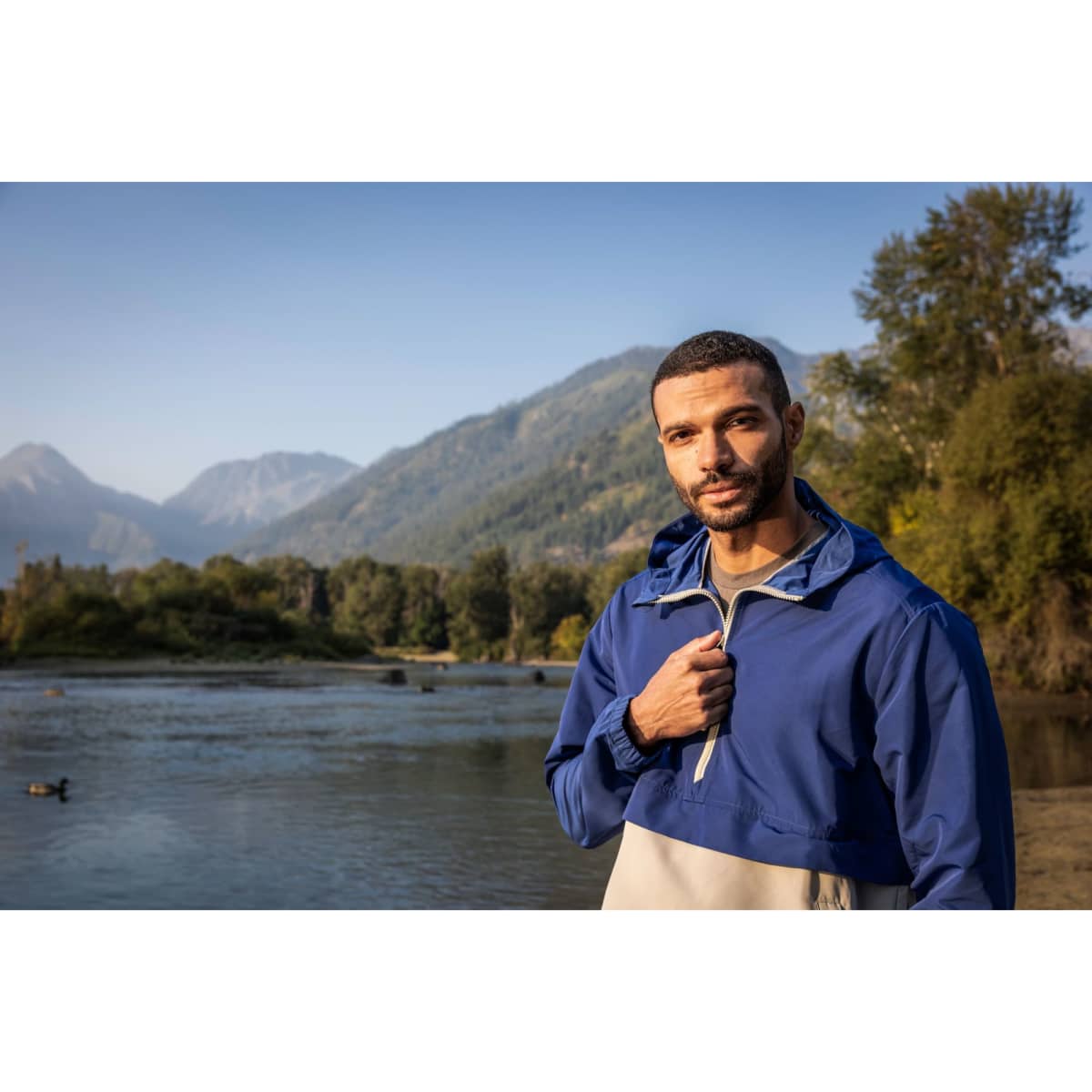 A man stands by a river wearing a blue and cream anorak jacket with a logo on the chest, surrounded by nature.