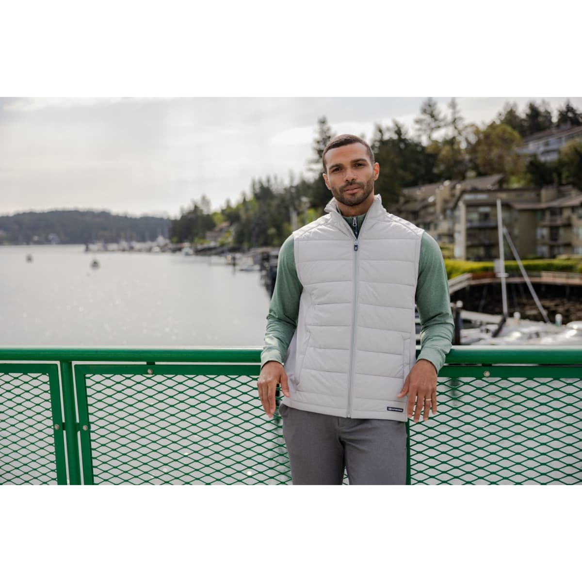 A man wearing a white eco hybrid vest over a green top, standing on a green railing with a waterfront background.