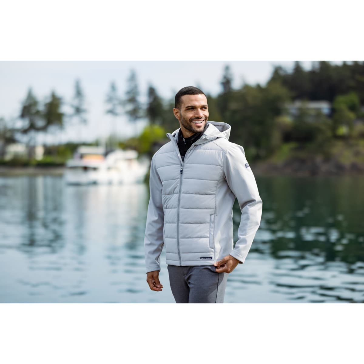 A man wearing a light grey hybrid jacket with a hood, smiling by the water, surrounded by trees and boats.