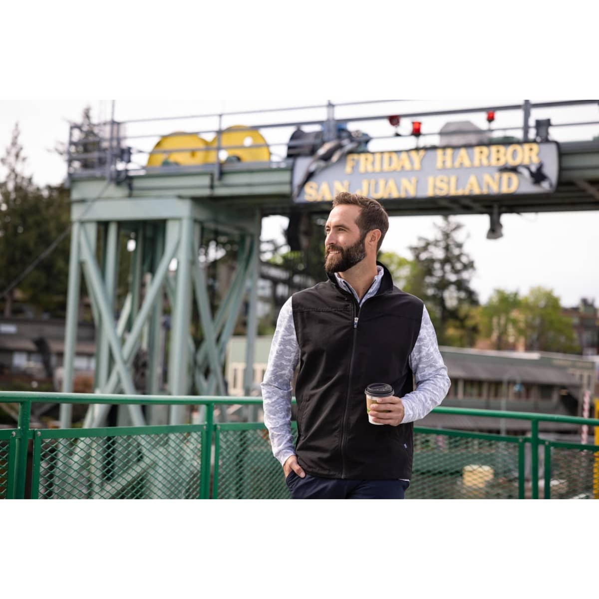 A man stands on a bridge wearing a black eco vest over a patterned shirt, holding a coffee cup.