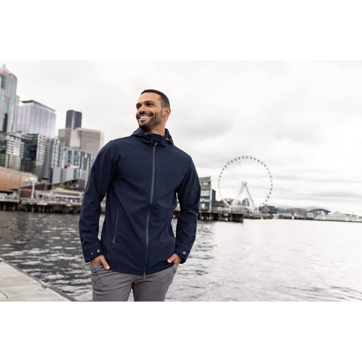 A man wearing a navy blue eco-friendly rain jacket stands by the water with a city skyline in the background.
