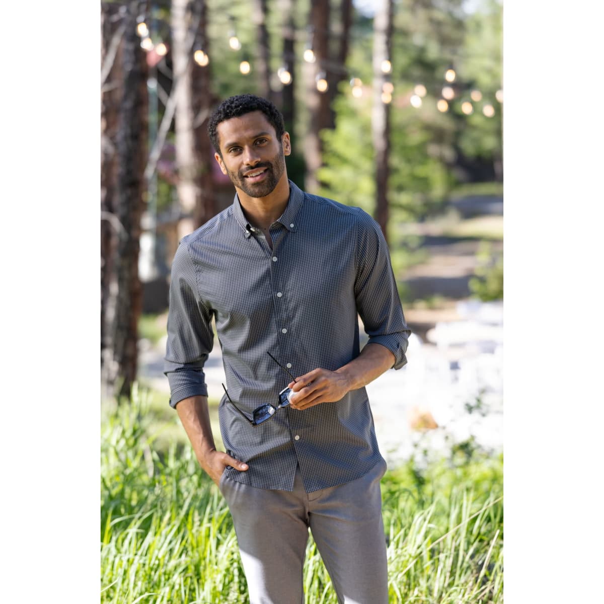 A man wearing a grey gingham shirt stands outdoors among greenery, holding sunglasses, with trees in the background.