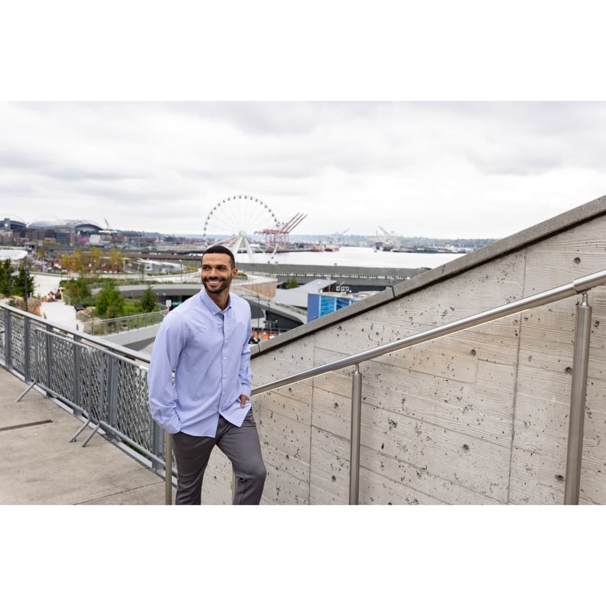 A man stands on stairs wearing a light blue pinstripe shirt and grey trousers, with a waterfront view in the background.