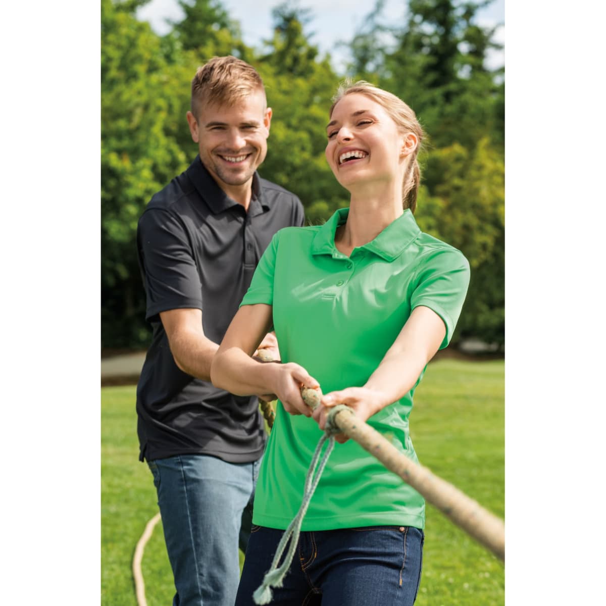 A man in a black polo and a woman in a green polo pull on a rope in a outdoor setting with trees in the background.