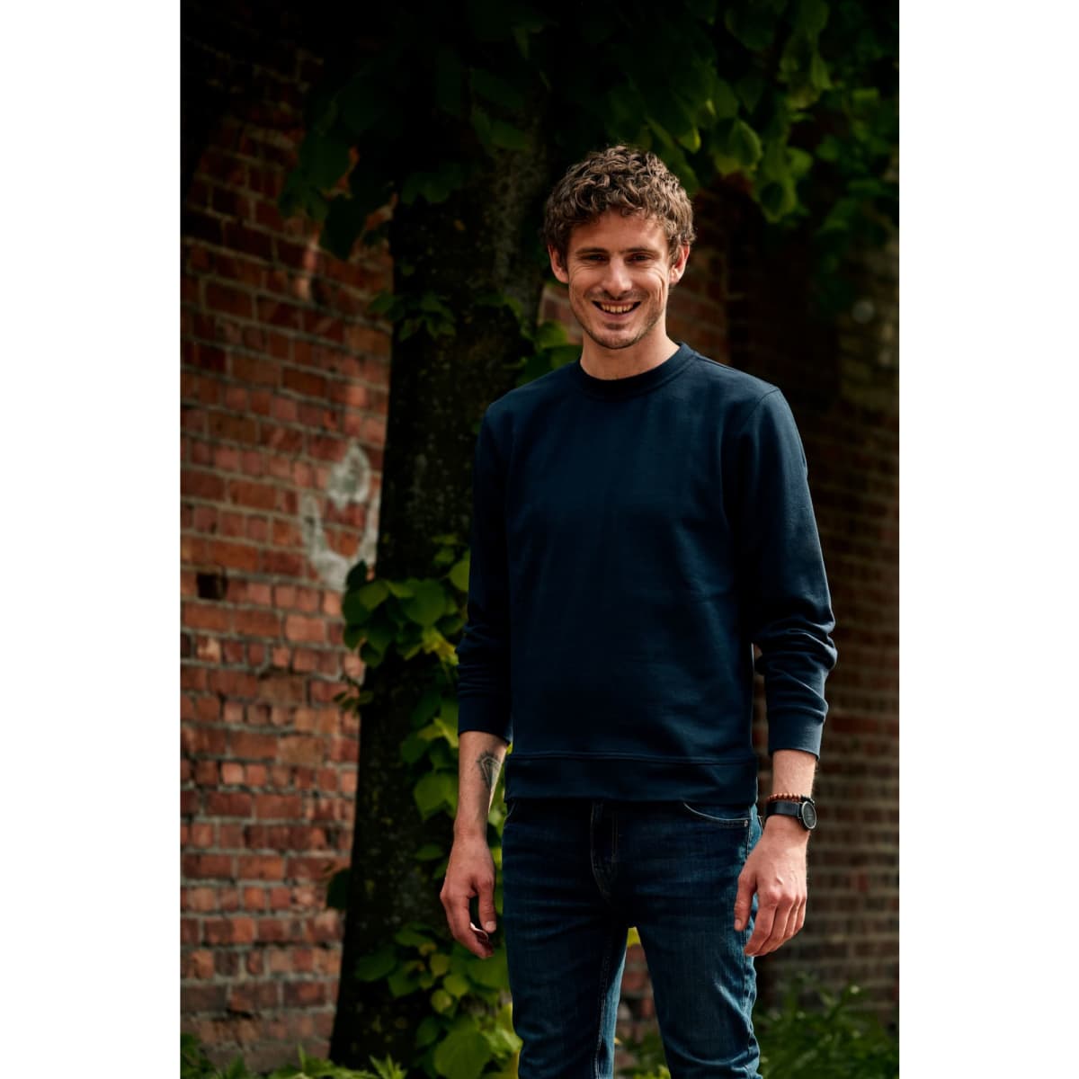 A man wearing a navy crewneck sweatshirt and blue jeans stands against a brick wall with greenery.
