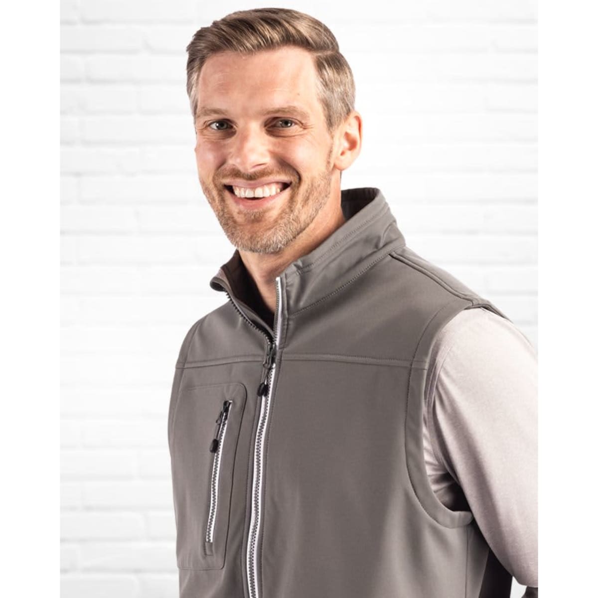 A grey men's softshell vest with a high collar and zip pockets, modelled by a smiling man against a white brick background.