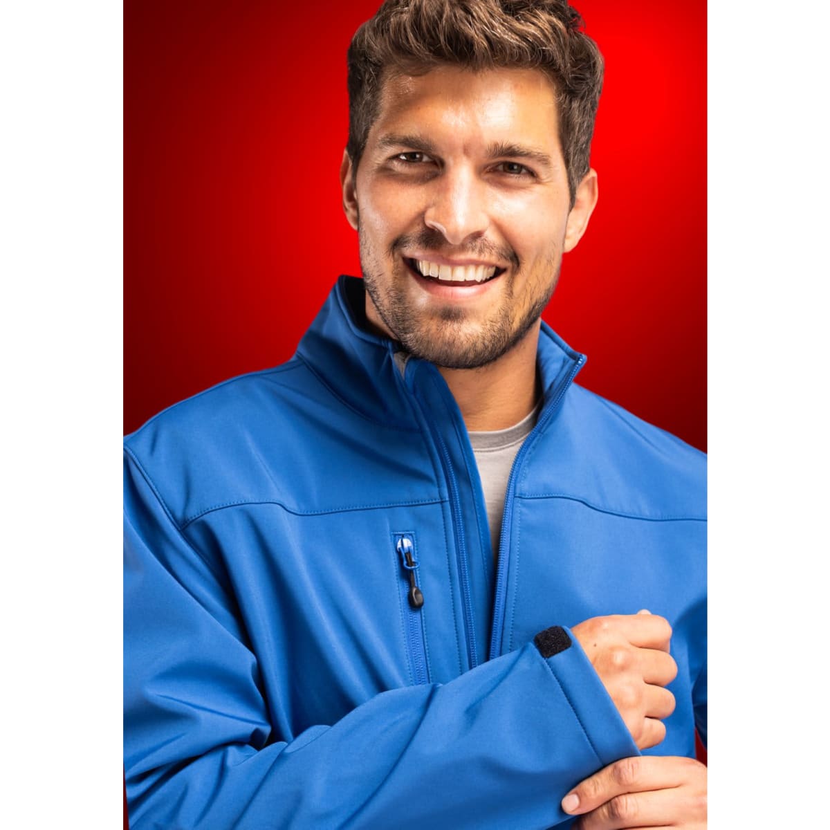 A man wearing a blue softshell jacket, smiling against a red background. The jacket has a logo on it.
