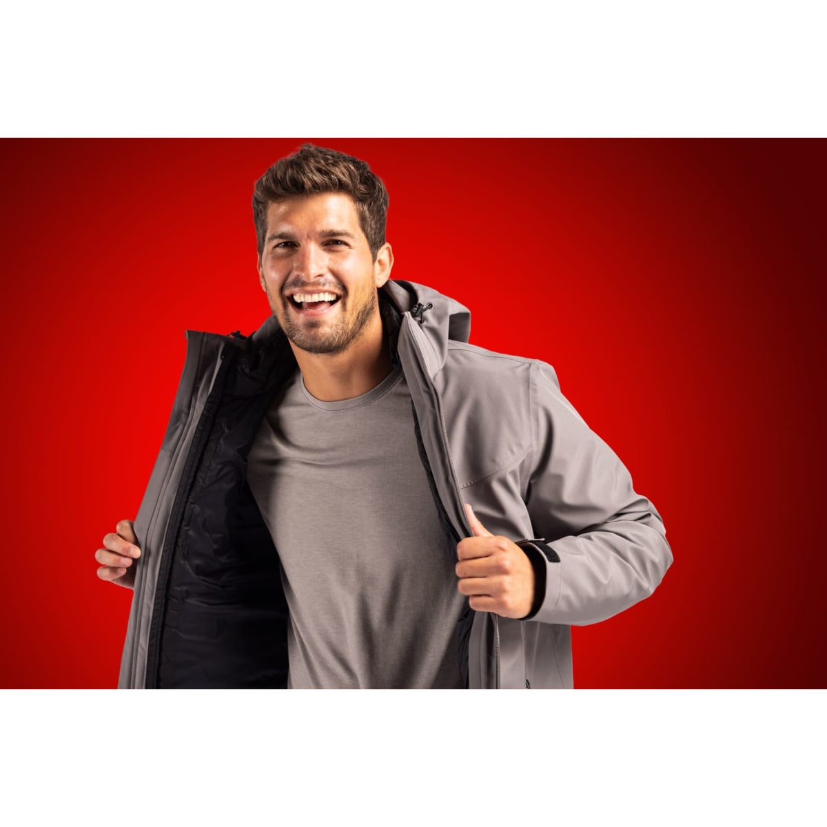 A man wearing a grey 3-in-1 parka jacket, smiling and opening the jacket, against a red background.