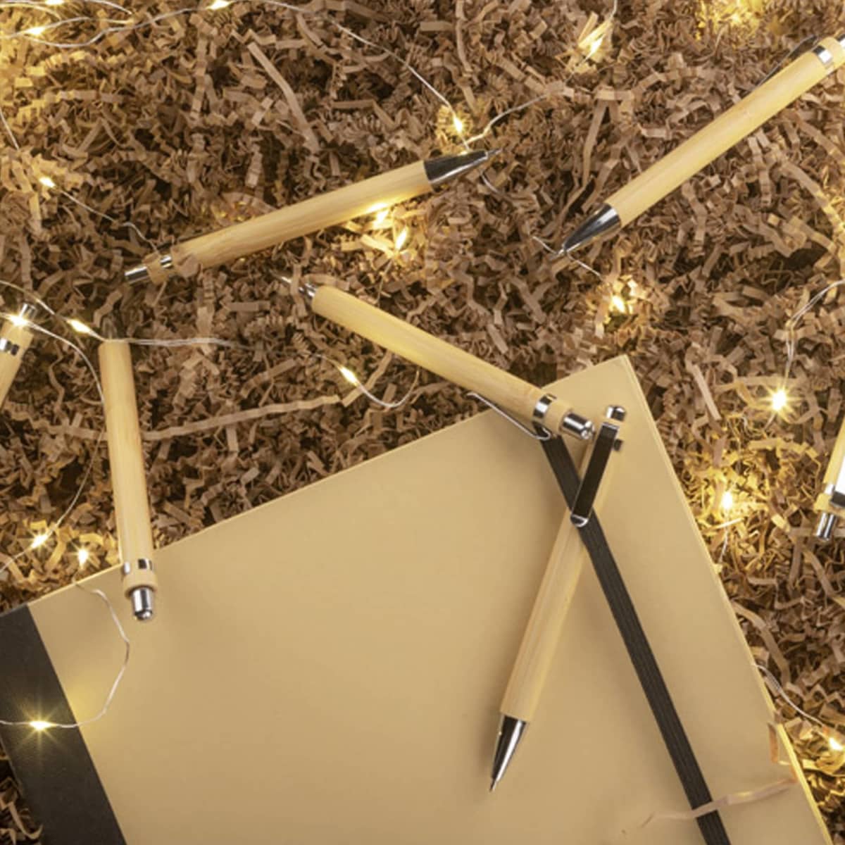 A collection of bamboo pens in natural colour scattered around a notebook and decorative lights on brown filler material.