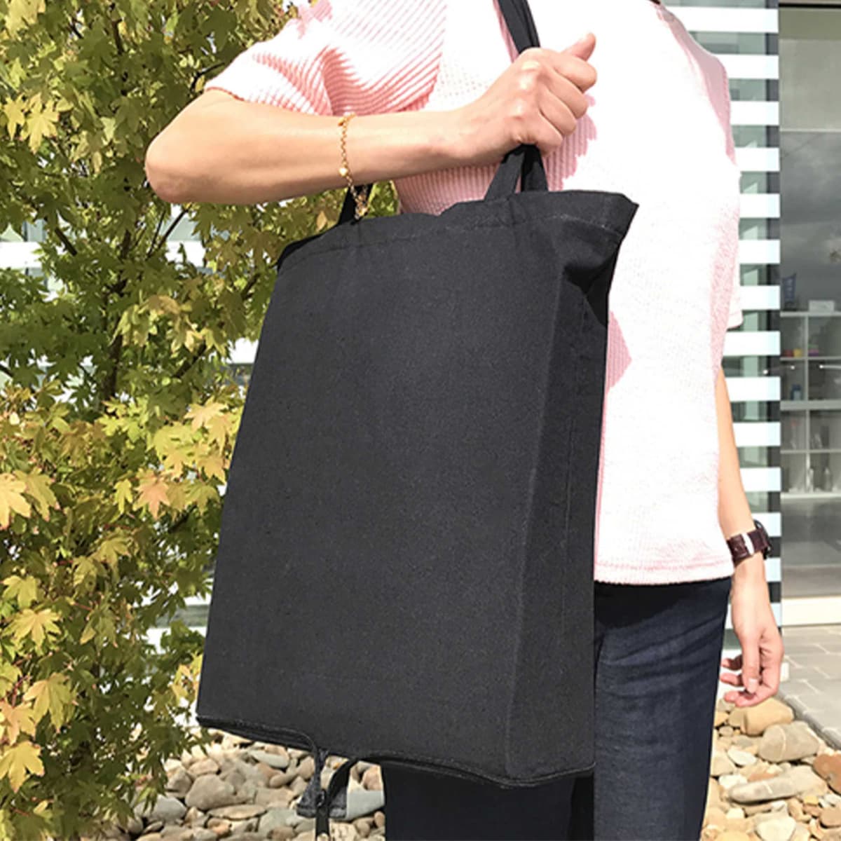 A person holds a black foldable tote bag against a backdrop of greenery. The bag has sturdy handles and a simple design.