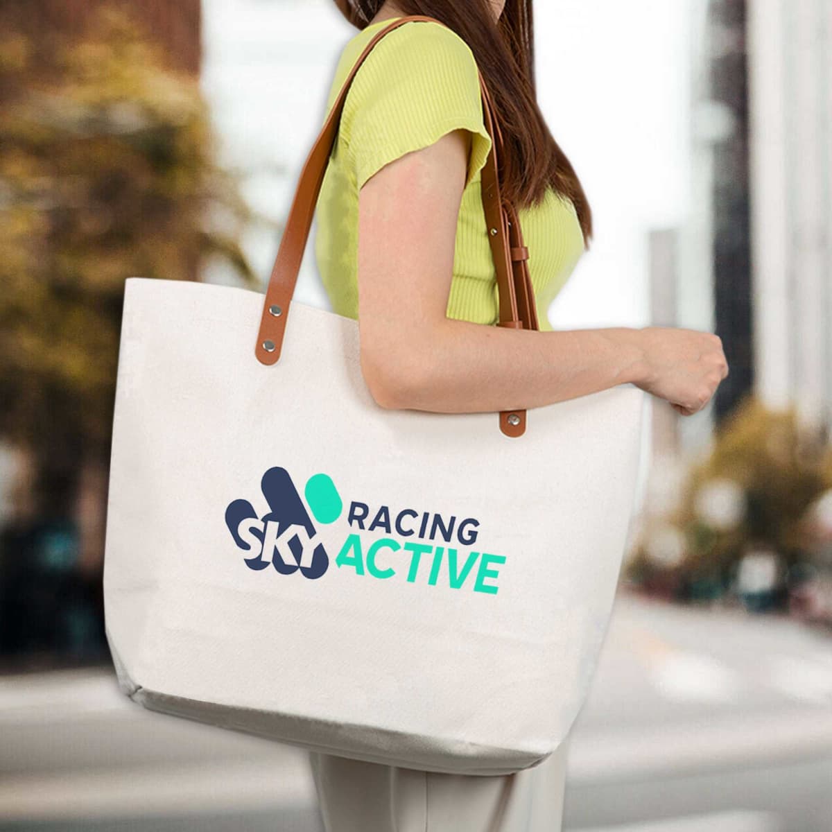 A tote bag in cream with brown handles, featuring a logo on the front, held by a woman outdoors.