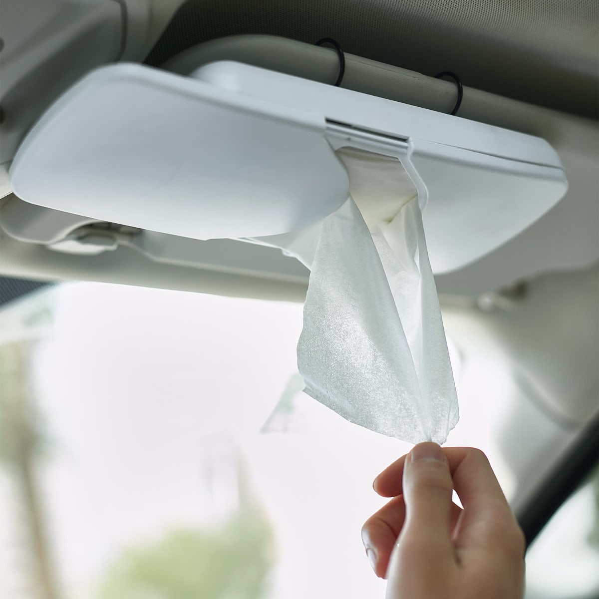 A white car sun visor tissue box with tissues being pulled out, mounted on a car's interior.