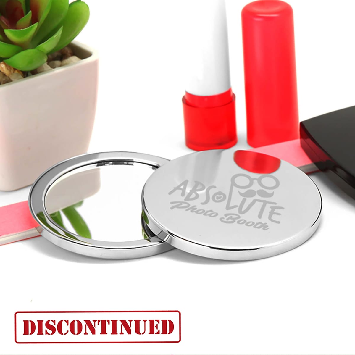 A compact mirror with a shiny silver finish, featuring a logo, placed next to a small plant and makeup items.