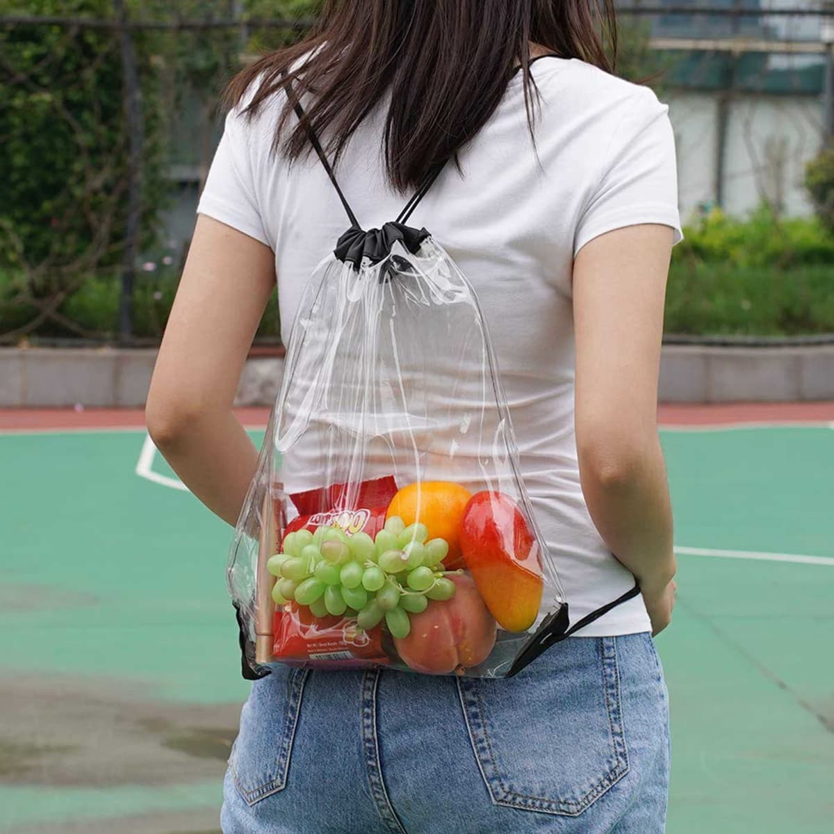 A clear drawstring backpack held by a person, filled with red apples, green grapes, and yellow oranges.
