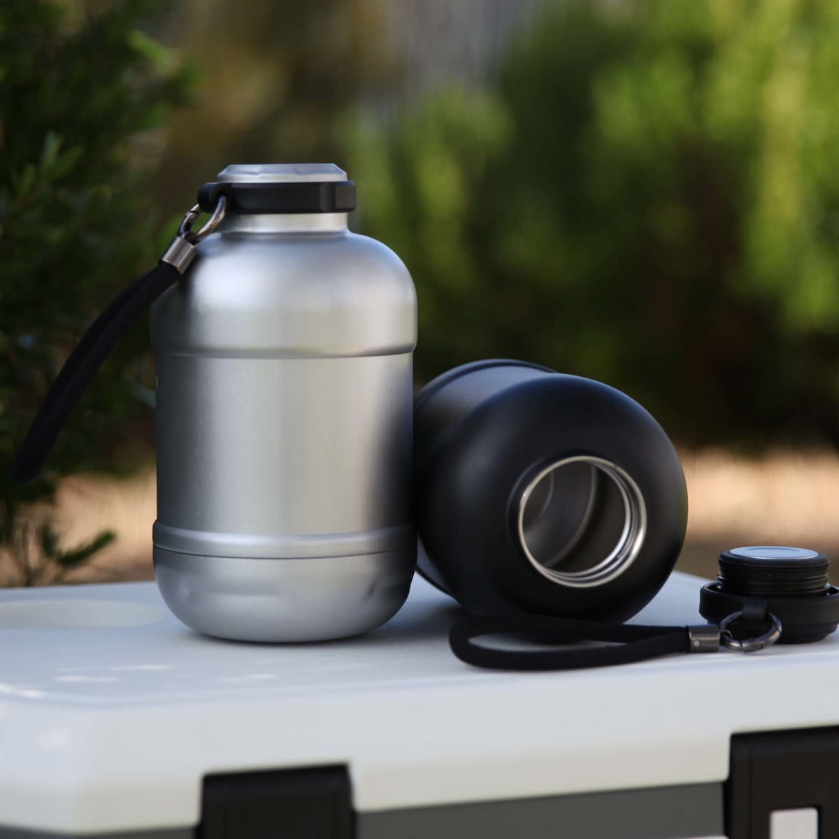 A drink bottle featuring a silver metallic finish and a black base, with a lid and strap, displayed on a cooler.