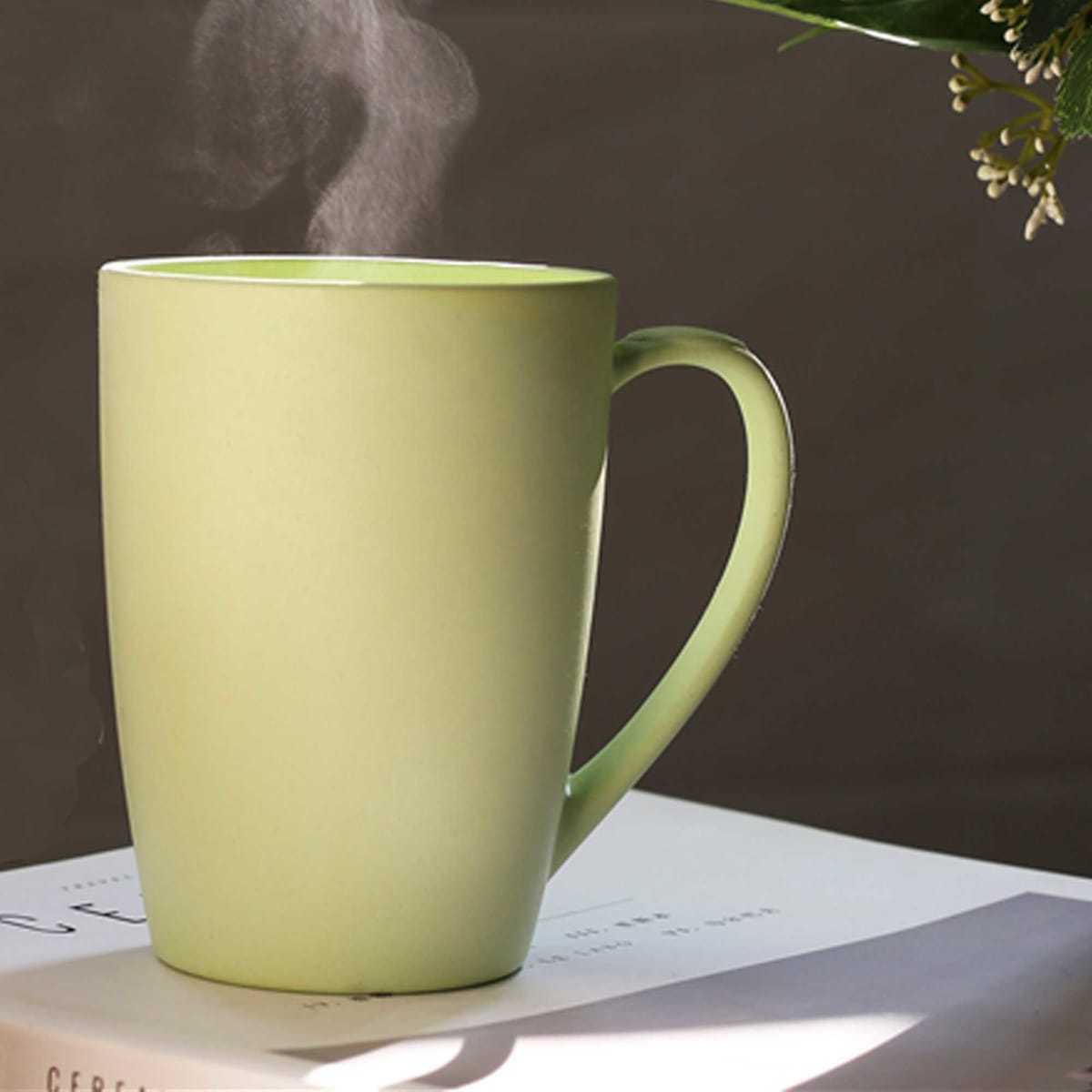 A green bamboo mug with a handle, emitting steam, resting on a white book.