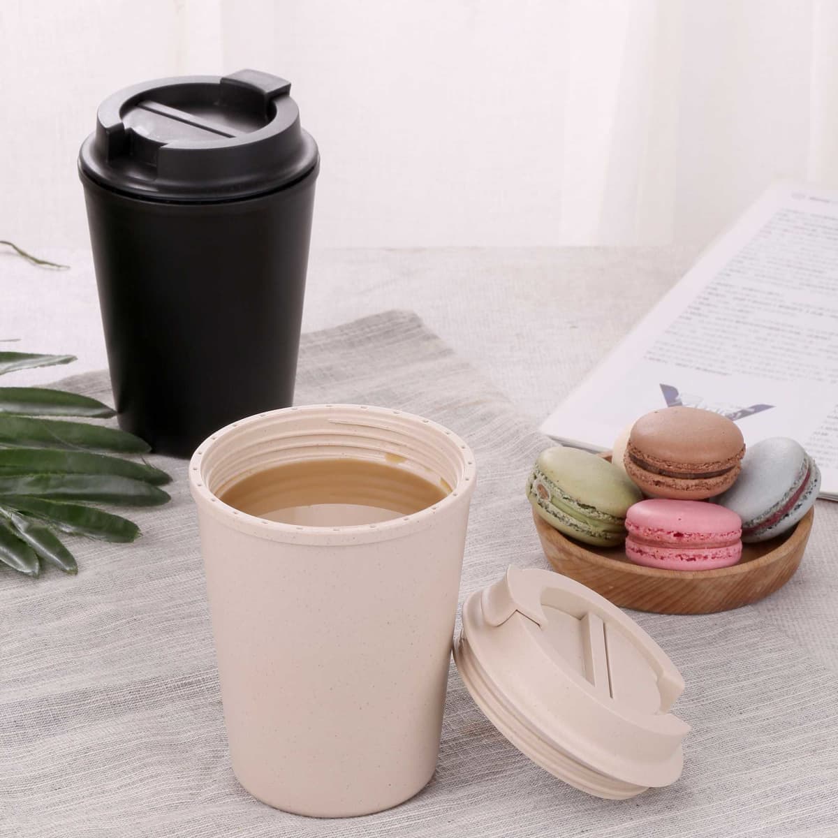 A drink bottle with a beige cup and a black cup, both featuring lids, alongside a plate of colourful macarons.