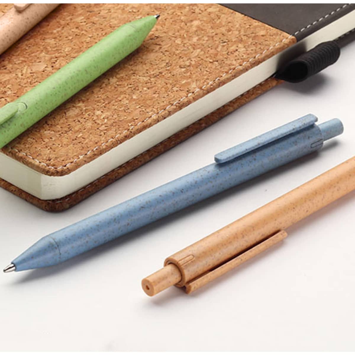 A collection of eco-friendly pens in green, blue, and tan, placed beside a cork notebook. The pens have a logo.