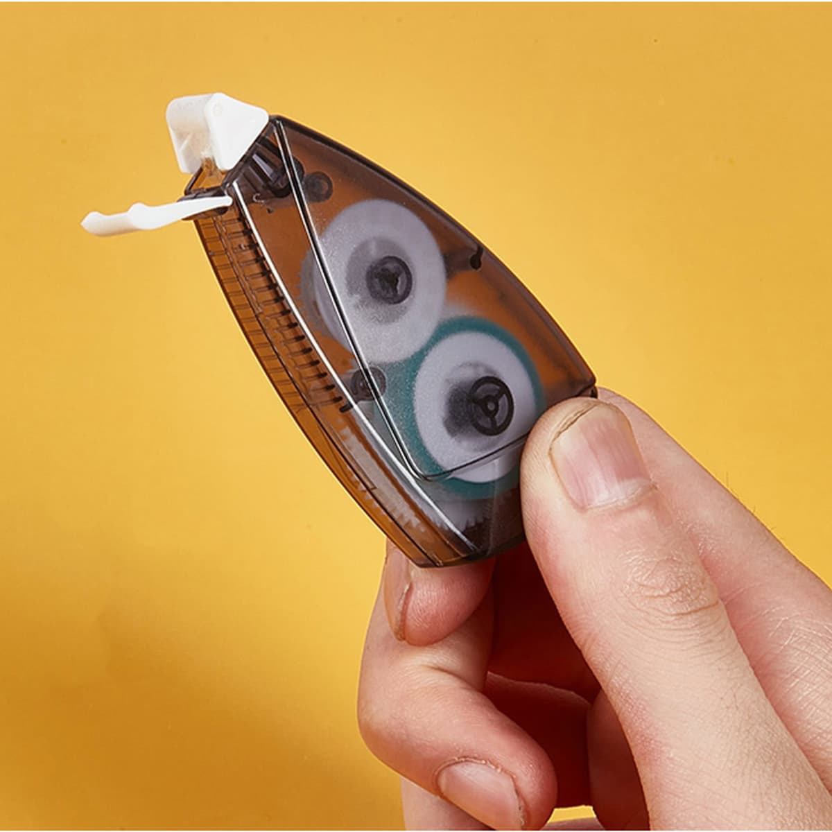 A hand holding a brown and transparent tape dispenser with a white tape inside, against a yellow background.