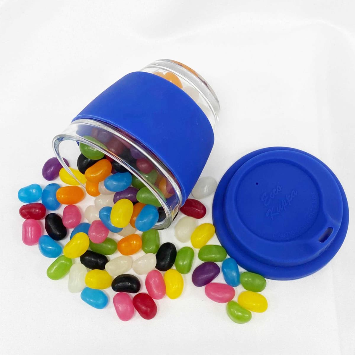 A glass coffee cup with a blue silicone sleeve, surrounded by an assortment of colourful jelly beans.