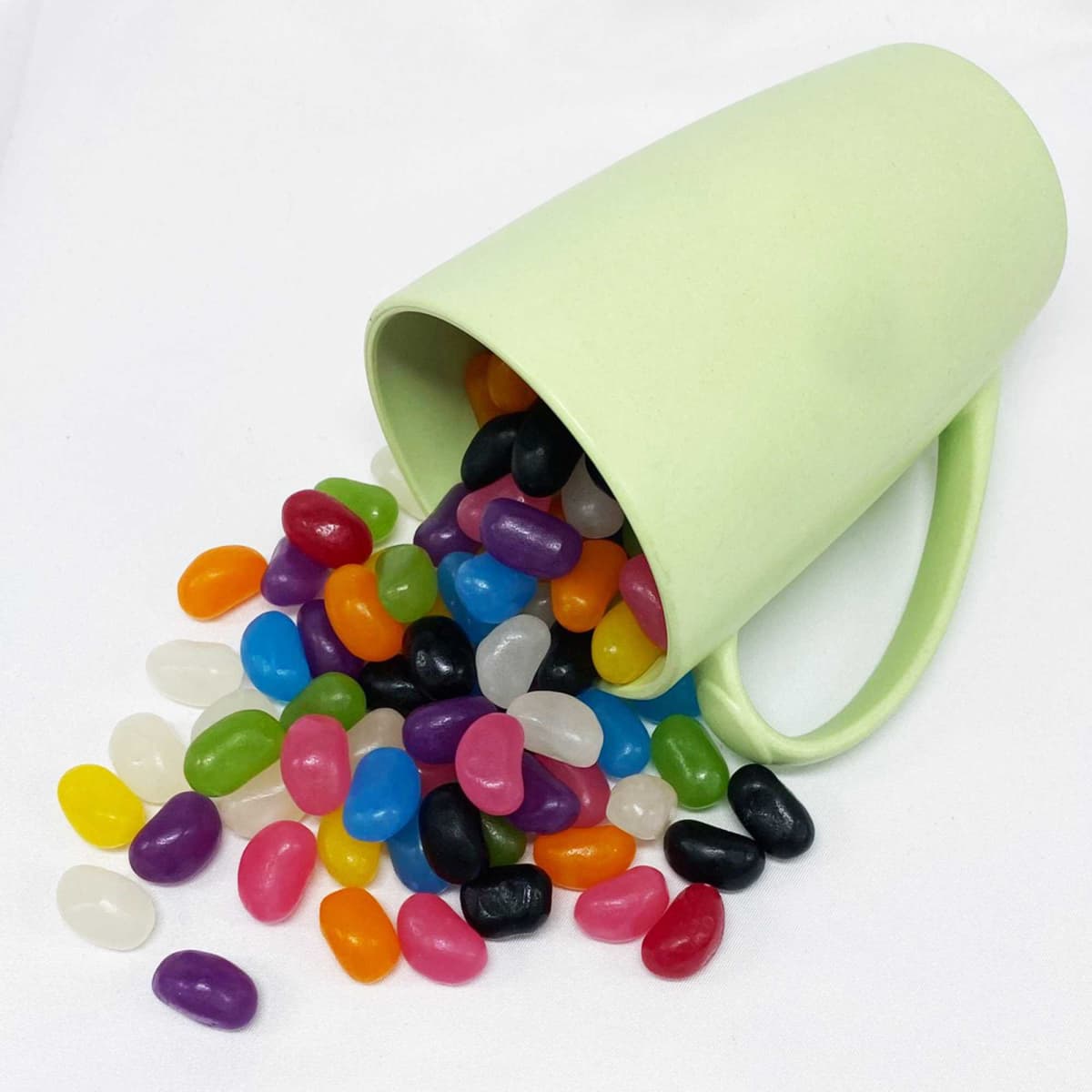 A green bamboo mug tipped over, spilling an assortment of colourful jelly beans on a white background.