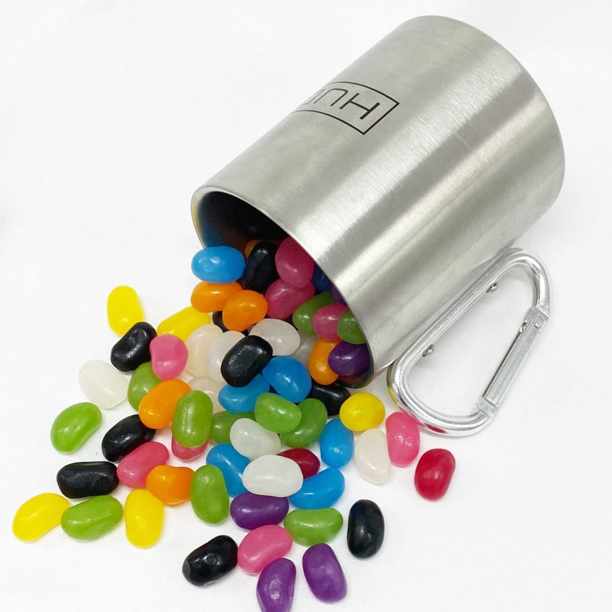 A stainless steel mug tipped over, spilling vibrant jelly beans in various colours onto a white surface.