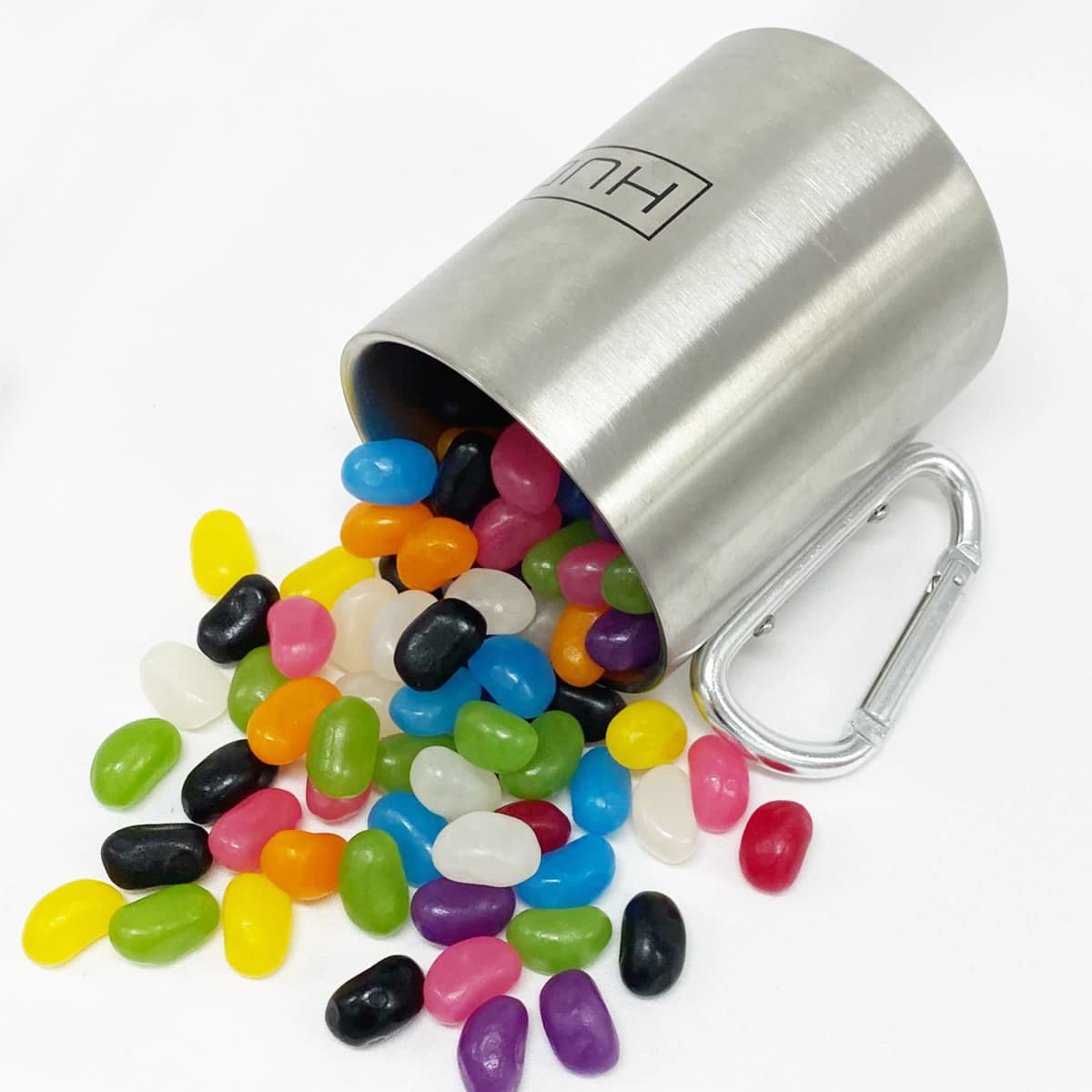 A stainless steel mug spills colourful jelly beans, including shades of yellow, pink, green, black, and white, with a carabiner.