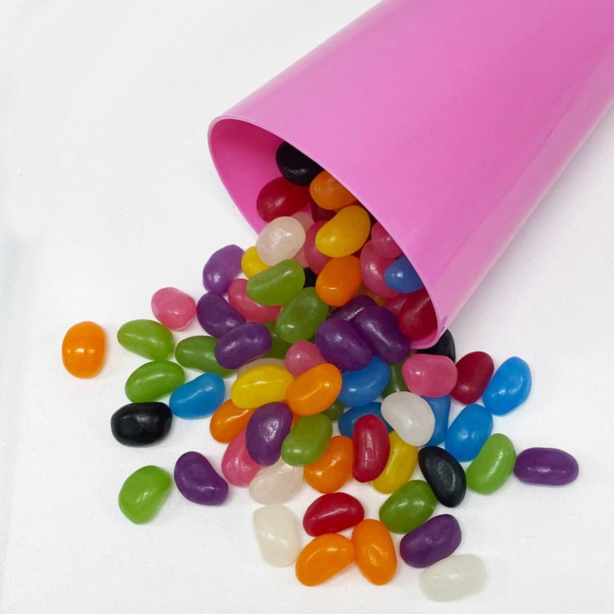 A pink party cup tipped over, spilling a mix of vibrant jelly beans in various colours on a white surface.